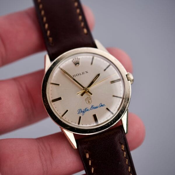 A hand holding a vintage Rolex Presentation Watch Ref. 7002 in 14k gold filled case, featuring a brown leather strap, silver dial, and classic Cal. 1520 movement.