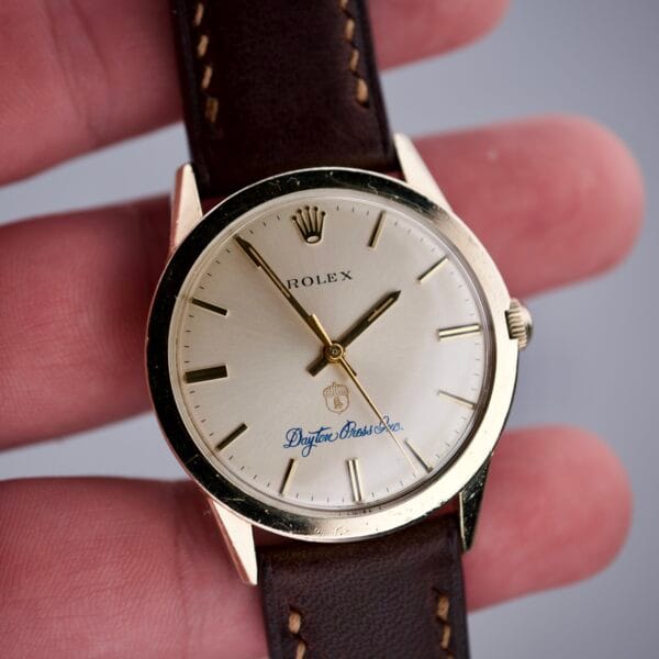 A hand holds a Rolex Presentation Watch Ref. 7002 with 14k gold filled case, brown leather strap, gold markers, and iconic Cal. 1520 movement.