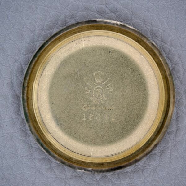 Top view of a round, metallic lid with embossed text and symbols on a textured gray surface, possibly from a Rolex Presentation Watch Ref. 7002 14k Gold Filled Cal. 1520.