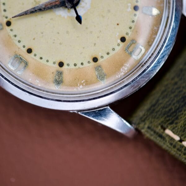 Close-up of a Bulova Wrist Alarm Manual Wind Cal. 11AERC featuring a worn beige dial, green leather strap, and stainless steel case.