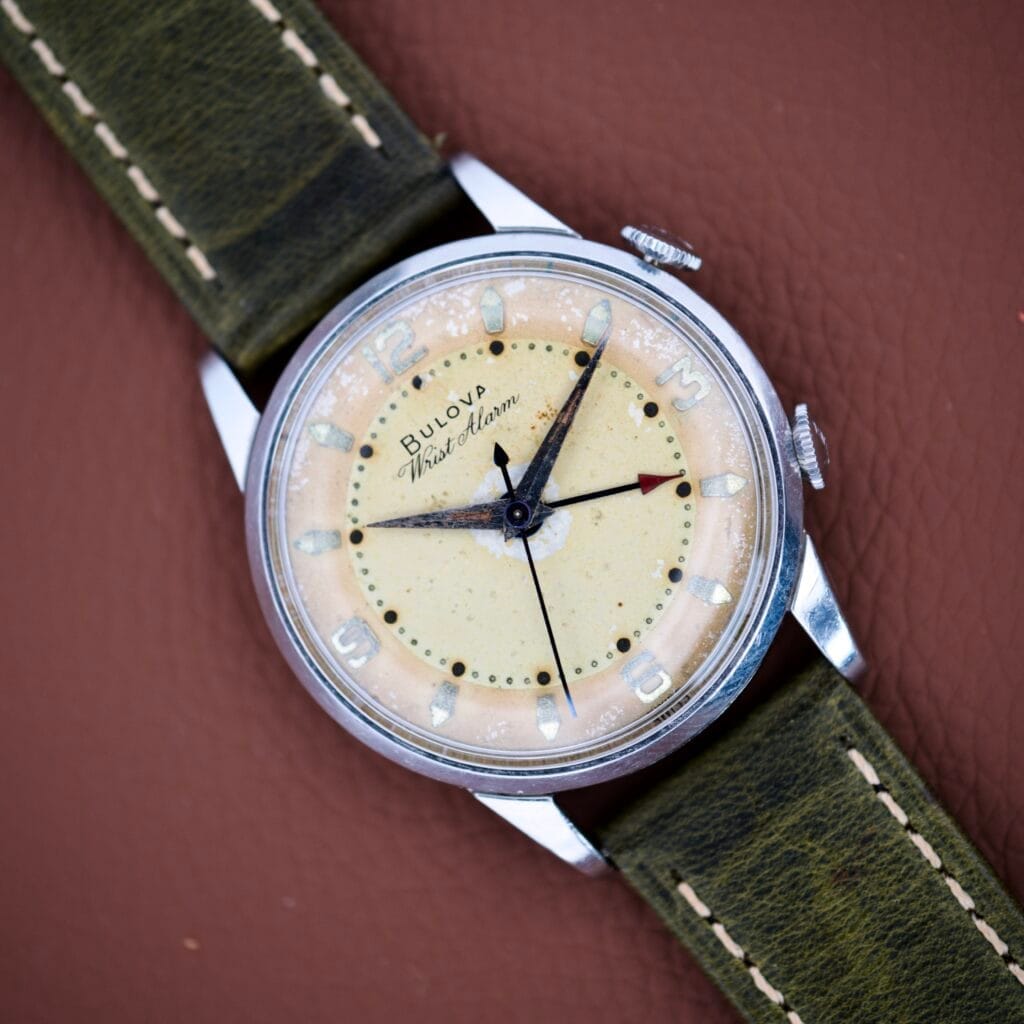 A Bulova Wrist Alarm Manual Wind Cal. 11AERC with a stainless steel case and green strap, displayed on a brown leather surface.