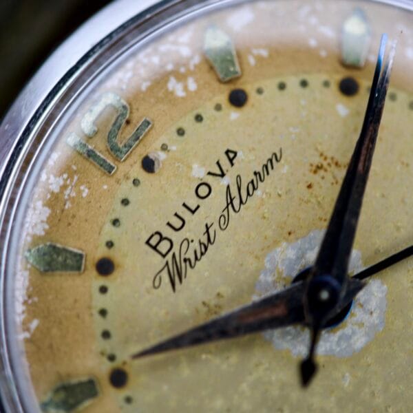 Close-up of a vintage Bulova Wrist Alarm Manual Wind Cal. 11AERC watch face with a weathered, yellowed dial and black hands, set in a classic stainless steel case.
