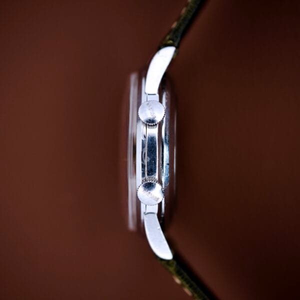 Side view of a Bulova Wrist Alarm Manual Wind Cal. 11AERC with a stainless steel case and two round buttons, shown against a brown background.