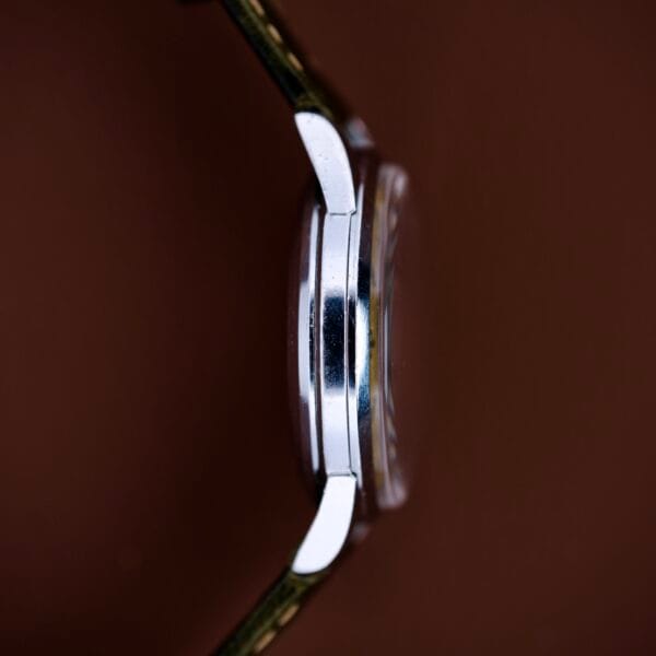 Side view of the Bulova Wrist Alarm Manual Wind Cal. 11AERC, featuring a sleek stainless steel case, displayed against a brown background.