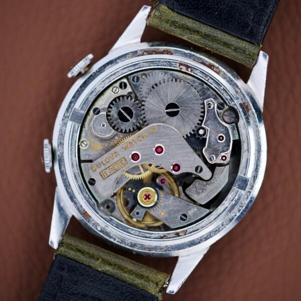 Close-up of a Bulova Wrist Alarm Manual Wind Cal. 11AERC movement, displaying visible gears and intricate mechanical parts within its classic stainless steel case.