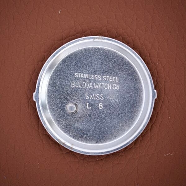 Bulova Wrist Alarm Manual Wind Cal. 11AERC stainless steel case back, marked SWISS L8, shown on a brown textured surface.