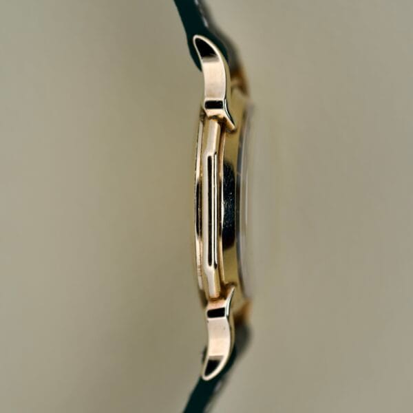 Side view of a Gruen Barclay "Spider" Ref. 335-723 gold wristwatch case with black leather strap, 21 Jewels Cal. 335, set against a plain beige background.