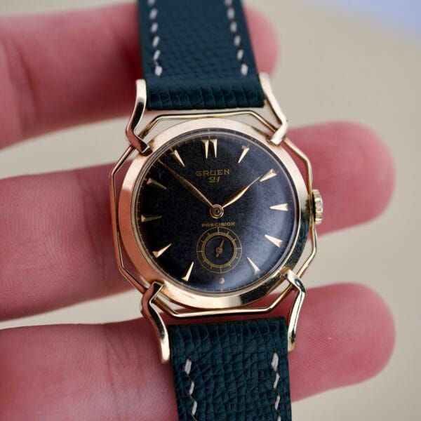 A hand holds a vintage Gruen Barclay "Spider" Ref. 335-723 wristwatch, featuring a gold case, black dial, leather strap, and 21 Jewels Cal. 335—an elegant classic.