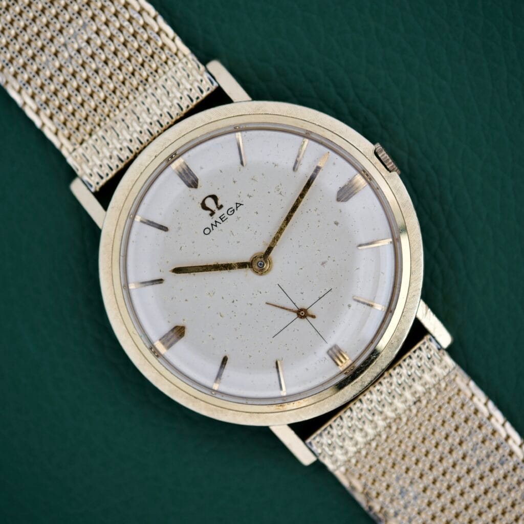 1962 Omega Ref. H6290 wristwatch with a cream dial, 14k gold filled case and hour markers, mesh metal band, and manual wind Cal. 510 movement—displayed on a green background for classic appeal.