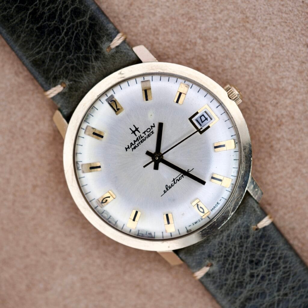 The Hamilton Masterpiece Electronic Date Ref. 981569 Cal. 702 features a silver dial, gold markers, black leather strap, Cal. 702 movement, and an electronic date window at 3 o’clock.