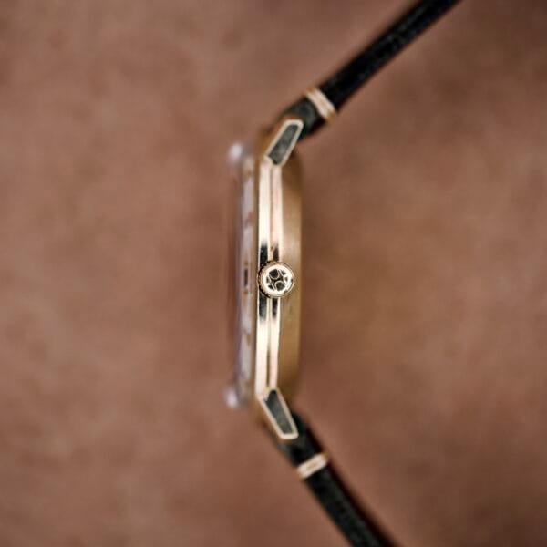 Side view of a Hamilton Masterpiece Electronic Date Ref. 981569 Cal. 702 gold wristwatch with a black leather strap, displayed against a brown background.