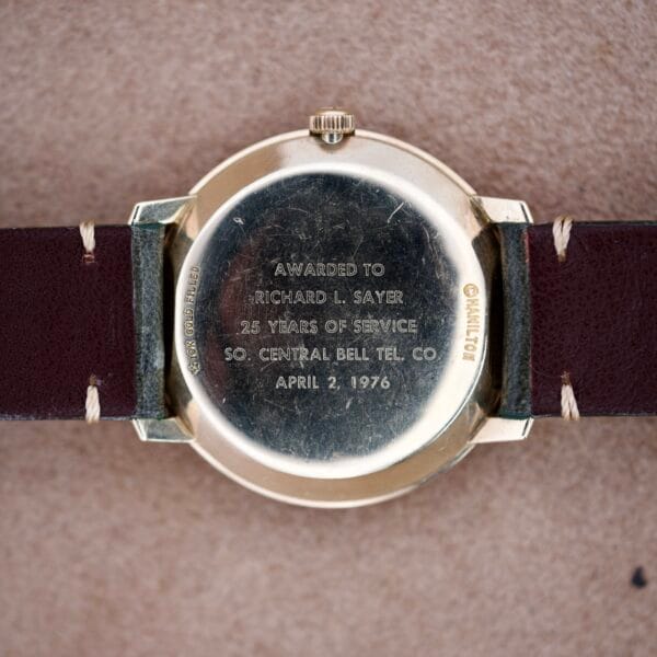 Hamilton Masterpiece Electronic Date Ref. 981569 Cal. 702 watch with a service award inscription, gold case, dark brown leather strap, and displayed on a beige surface.