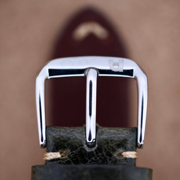 Close-up of a silver belt buckle on dark leather, echoing the refined style of the Hamilton Masterpiece Electronic Date Ref. 981569 Cal. 702, set against a blurred brown background.
