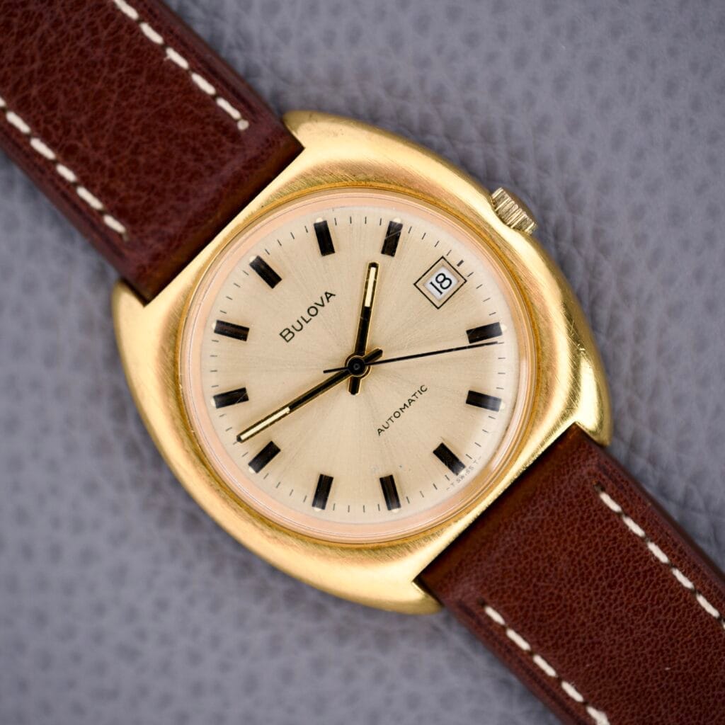 Bulova Cushion Case Automatic Ref. 7313-2 Cal. 11AOACD wristwatch with a gold case and brown leather strap displayed on a gray textured background.