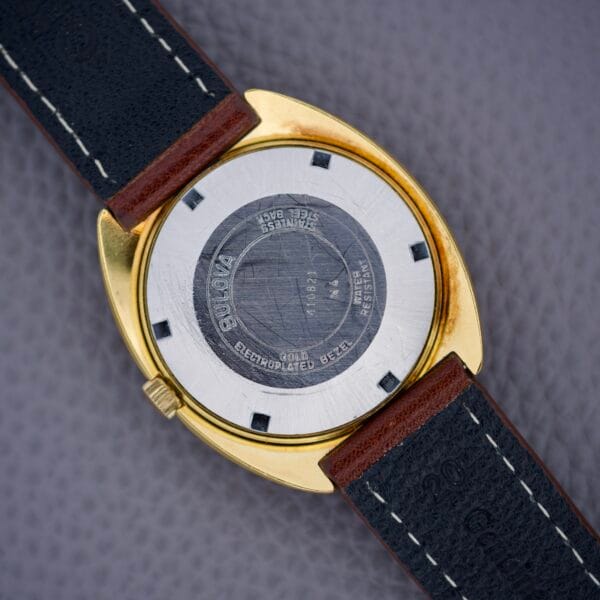 Back of a Bulova Cushion Case Automatic Ref. 7313-2 Cal. 11AOACD with a gold-tone case and black leather strap, shown on a textured gray surface.