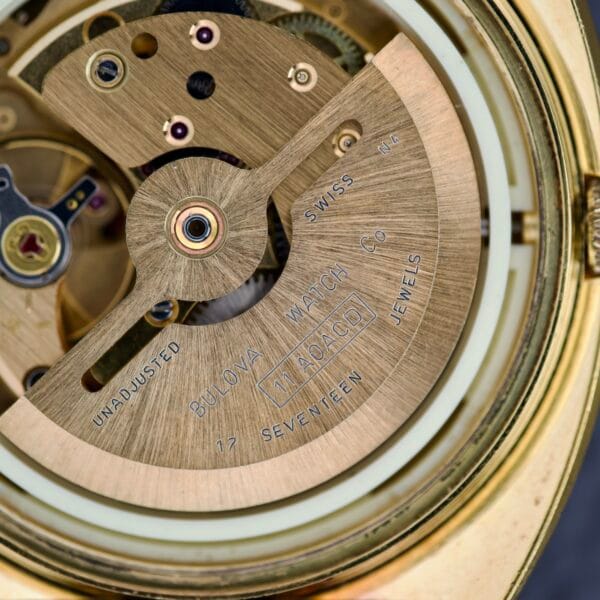 Close-up of a Bulova Cushion Case Automatic Ref. 7313-2 Cal. 11AOACD watch movement, showing gears and engraved text on the gold rotor inside its iconic cushion case.