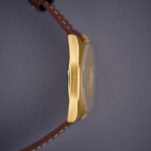 Side view of a Bulova Cushion Case Automatic Ref. 7313-2 Cal. 11AOACD gold wristwatch with a brown leather strap on a textured gray background.