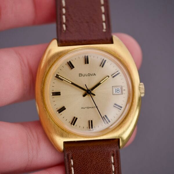 The Bulova Cushion Case Automatic Ref. 7313-2 Cal. 11AOACD features a gold case, brown leather strap, date display, and black hour markers, powered by the Cal. 11AOACD movement.