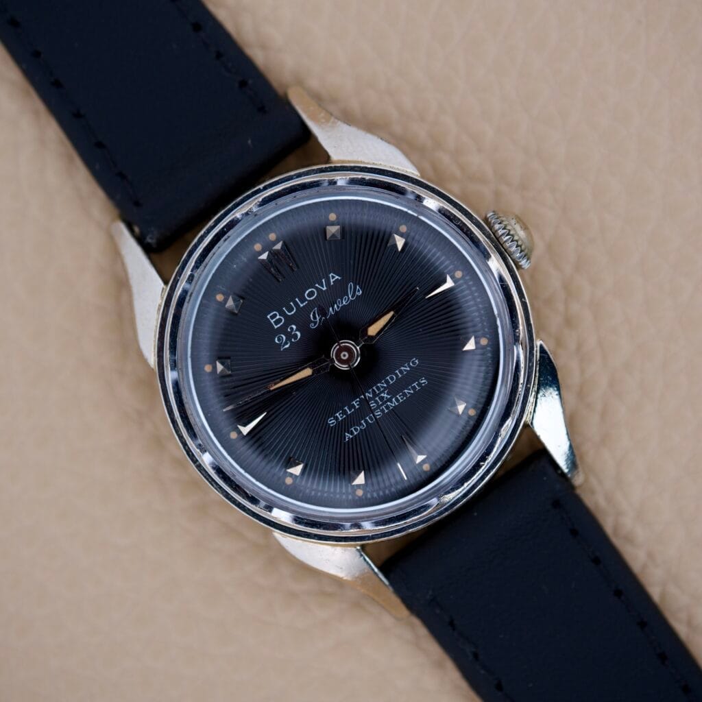 A Bulova 23 Jewels Automatic Six Adjustments Cal. 10BPAC wristwatch with a black strap and black dial is displayed on a beige textured surface.