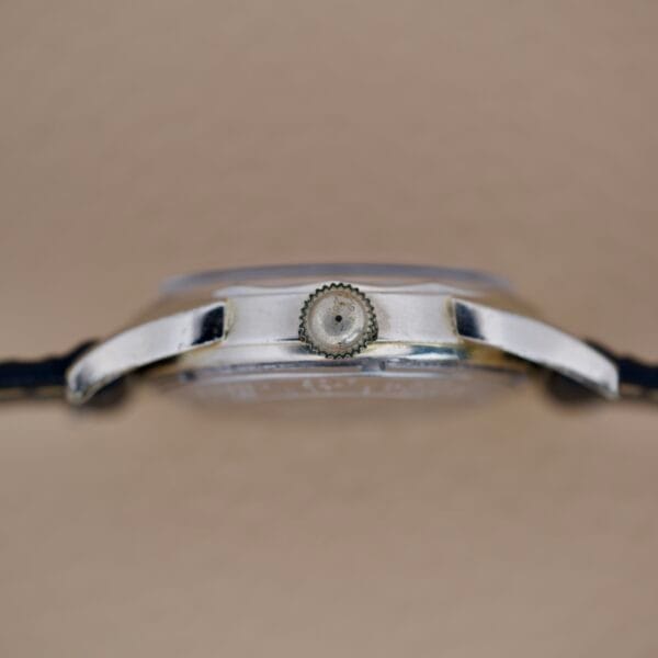 Side view of a Bulova 23 Jewels Automatic Six Adjustments Cal. 10BPAC wristwatch with black strap, visible crown, and beige background.