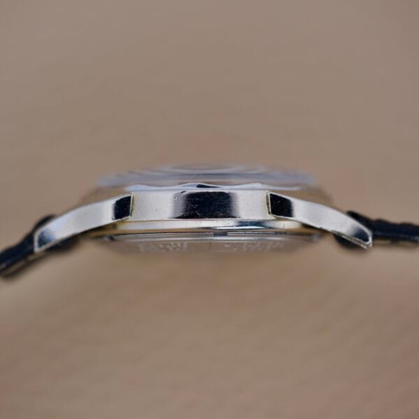 Side view of the Bulova 23 Jewels Automatic Six Adjustments Cal. 10BPAC wristwatch with a black strap, showcasing its thin case against a beige background.