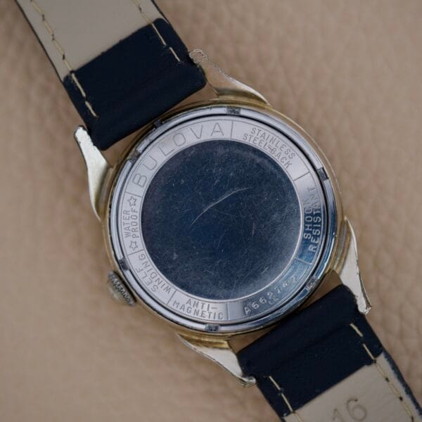 The back of a Bulova 23 Jewels Automatic Six Adjustments Cal. 10BPAC wristwatch with a black leather strap is shown on a beige surface, highlighting its engraved details.