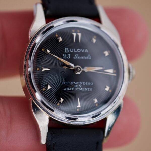 A close-up of a Bulova 23 Jewels Automatic Six Adjustments Cal. 10BPAC wristwatch with a black dial, silver case, and black leather strap.