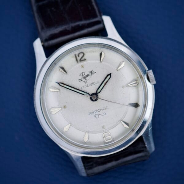A Piguette Antioch wristwatch with a white dial, black leather strap, and stainless steel case on a blue surface, featuring manual wind movement with 17 jewels for smooth performance.