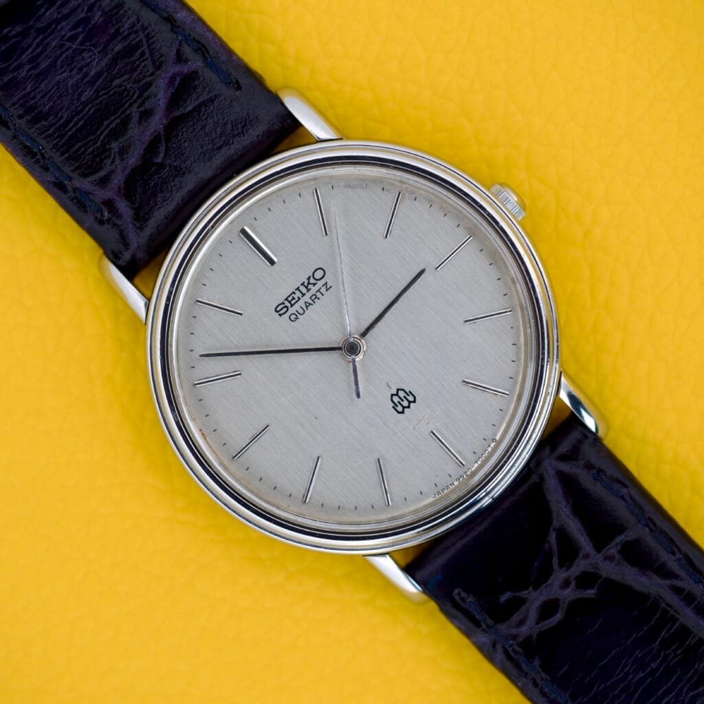 A Seiko Twin Quartz Ref. 9920-7000T Thin Stainless Steel wristwatch with a black leather strap rests on a yellow textured surface.