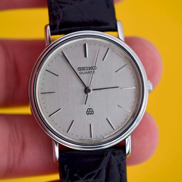 A hand holds the Seiko Twin Quartz Ref. 9920-7000T Thin Stainless Steel wristwatch with a black leather strap, set against a yellow background.
