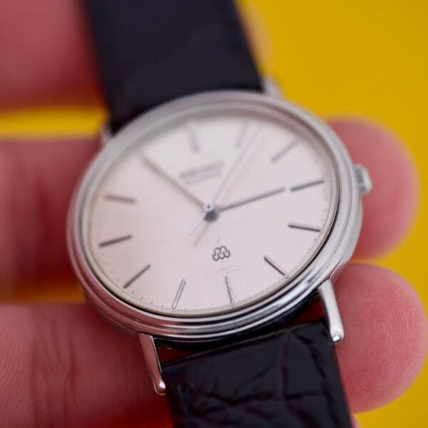 A hand holds a Seiko Twin Quartz Ref. 9920-7000T Thin Stainless Steel analog wristwatch with a black leather strap, set against a yellow background.