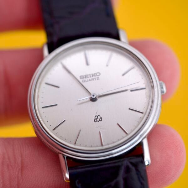 A hand holds a Seiko Twin Quartz Ref. 9920-7000T Thin Stainless Steel watch with a silver face and black leather strap against a yellow background.
