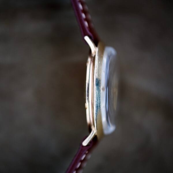 Close-up side view of Finley's Nivada Grenchen Automatic White Dial wristwatch with a gold case and brown leather strap, set against a blurred background.