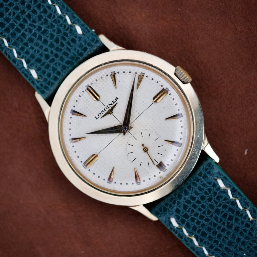 A Longines Waffle Dial Ref. 1064 wristwatch with a 10k Gold Filled case, white waffle dial, and green textured strap is displayed against a brown background. Powered by Cal. 23Z movement.