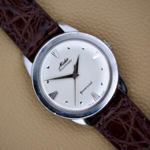 The Mido Multifort Powerwind White Dial Automatic Cal. 917P rests on a brown leather strap atop a beige textured surface.