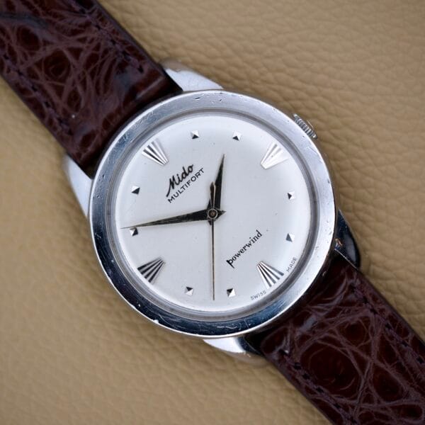 The Mido Multifort Powerwind White Dial Automatic Cal. 917P rests on a brown leather strap atop a beige textured surface.