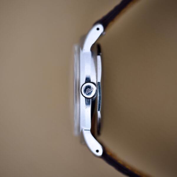 Side view of a silver Mido Multifort Powerwind White Dial Automatic Cal. 917P wristwatch with a brown leather strap, set against a beige background.