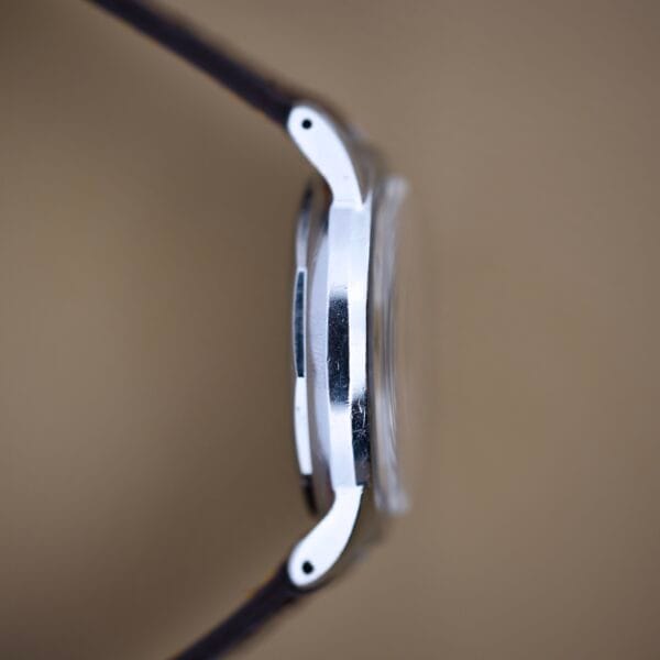 Side view of a Mido Multifort Powerwind wristwatch with white dial and leather strap, featuring Automatic Cal. 917P, set against a blurred brown background.