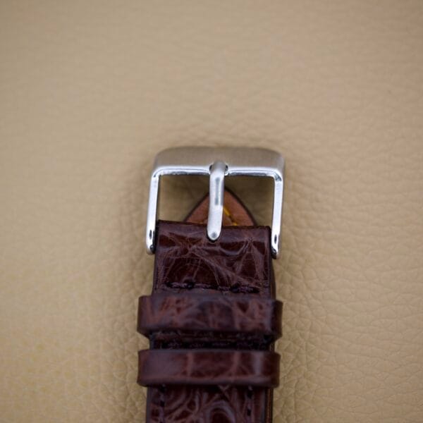 The Mido Multifort Powerwind White Dial Automatic Cal. 917P features a brown leather strap with a silver buckle, inspired by classic Multifort style, displayed on a beige textured surface.