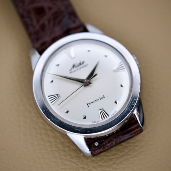 The Mido Multifort Powerwind White Dial Automatic Cal. 917P wristwatch features a white dial, silver case, and brown leather strap, elegantly displayed on a tan surface.