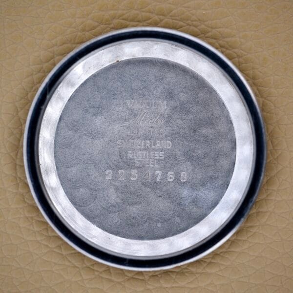 The round metal case back of a Mido Multifort Powerwind White Dial Automatic Cal. 917P, engraved with numbers, sits on a textured tan surface.