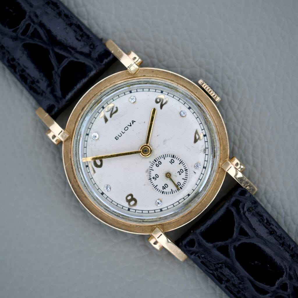 Bulova Princeton 10k gold filled case wristwatch with diamond dial (Cal. 10AK) features a black leather strap and a white face with gold accents, displayed on a gray textured surface for timeless elegance.