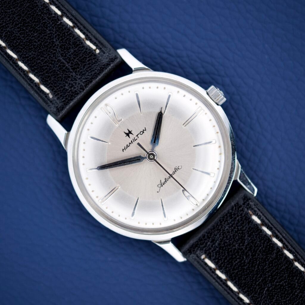 A Hamilton Accumatic X Sunburst Dial Stainless Steel Cal. 667 automatic watch with a black leather strap rests on a blue textured surface.