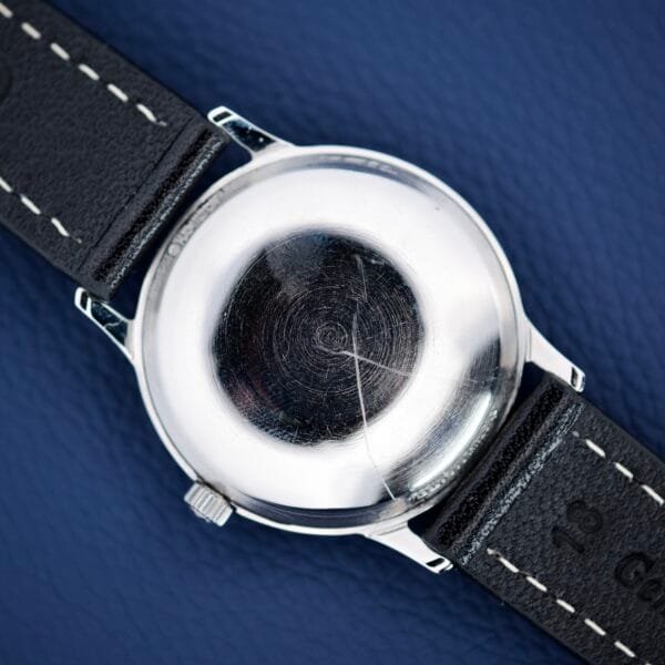 A close-up of a Hamilton Accumatic X Sunburst Dial Stainless Steel Cal. 667’s scratched stainless steel case back and black leather strap on a dark blue surface.