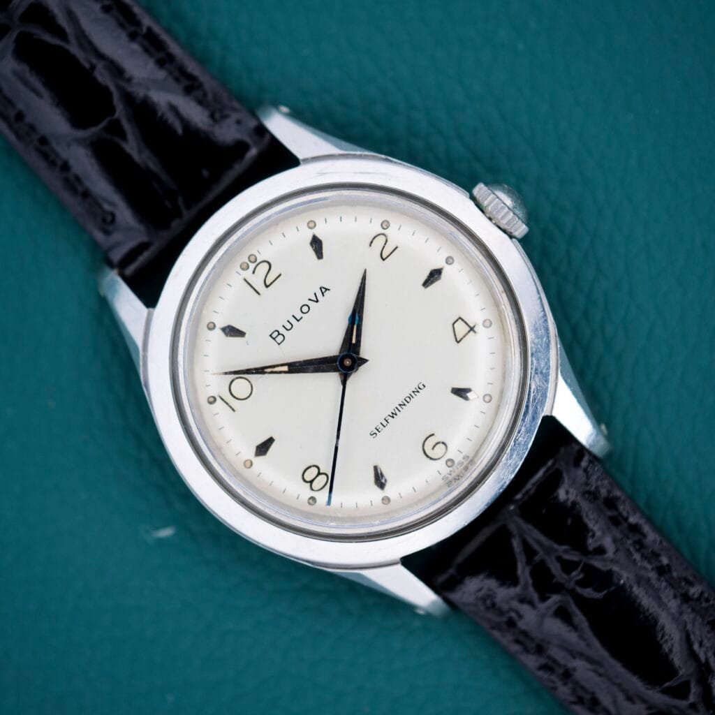 A 1955 Bulova Automatic Stainless Steel Cal. 11ACAC wristwatch with a black leather strap rests on a teal textured surface.