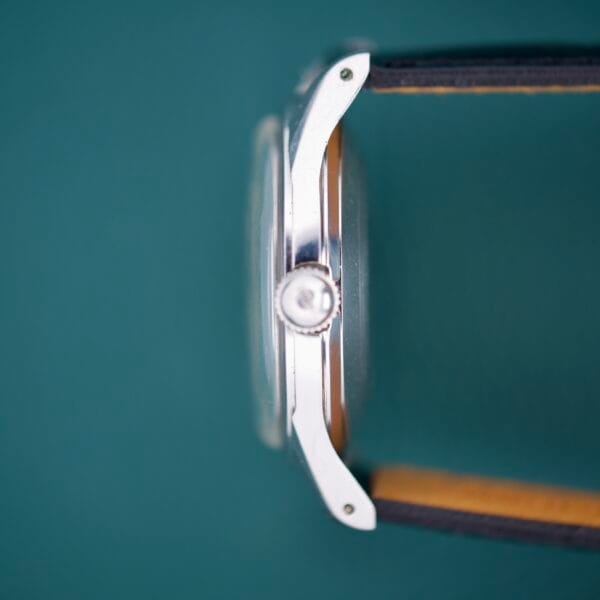 Side view of the 1955 Bulova Automatic Stainless Steel Cal. 11ACAC wristwatch with a leather strap, showing the crown and silver case against a green background.