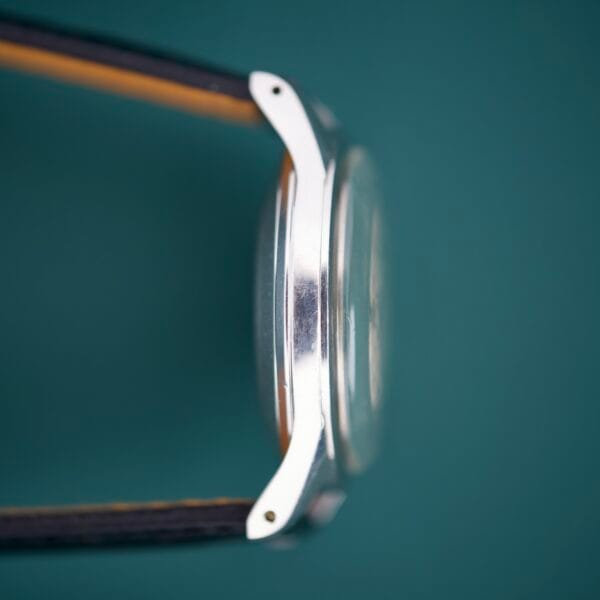 Side view of the 1955 Bulova Automatic Stainless Steel Cal. 11ACAC with a black and brown strap, set against a teal background.