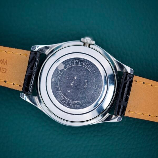 The back of a 1955 Bulova Automatic Stainless Steel Cal. 11ACAC wristwatch with a leather strap shows engraved text—Cal. 11ACAC and Stainless Steel—on the metal case.