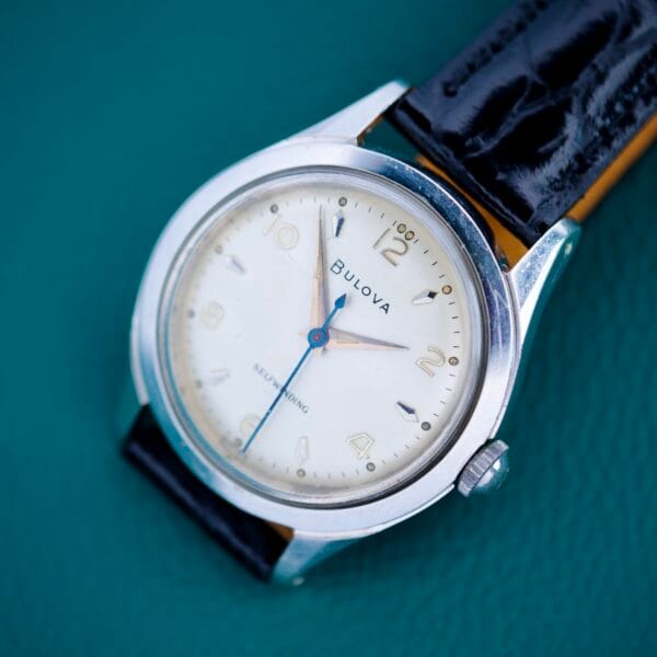 A 1955 Bulova Automatic Stainless Steel Cal. 11ACAC wristwatch with a black leather strap rests elegantly on a teal surface.