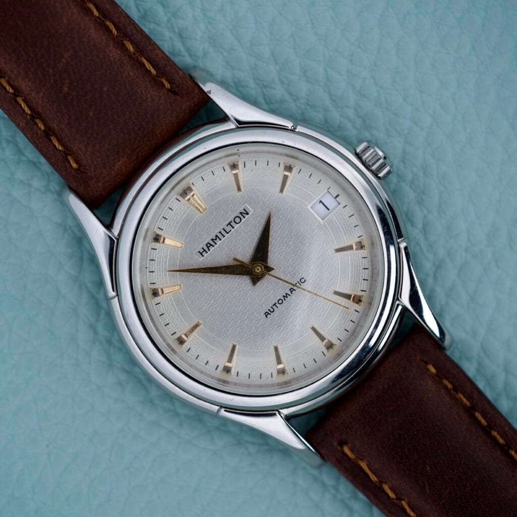 A Hamilton Automatic Date Ref. 6239 with 25 jewels and a brown leather strap rests on a light blue textured surface.